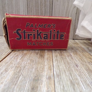 May include: A vintage red matchbox with the text "PALMER'S Strikalite MATCHES" in black and gold lettering. The matchbox is slightly worn and sits on a wooden surface.