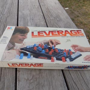 May include: A vintage board game box titled "LEVERAGE" by Milton Bradley. The box features a game board with red, blue, and white game pieces. The box art shows people playing the game. The game is for ages 10 and up, for 2 players.