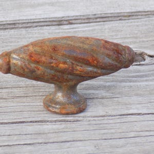 May include: A cabinet knob featuring a horizontal, elongated, and twisted cylindrical shape with a weathered, rusted brown and grey patina. The hardware includes a circular base that supports the central handle, showcasing a textured, aged metal finish.
