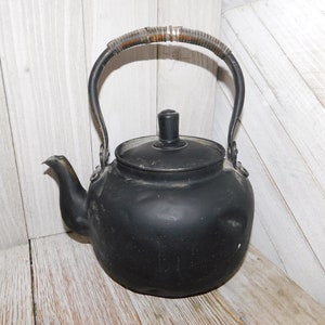 May include: A black metal teapot with a wooden handle. The teapot has a rounded body and a spout on the side. The teapot is sitting on a white wooden surface.