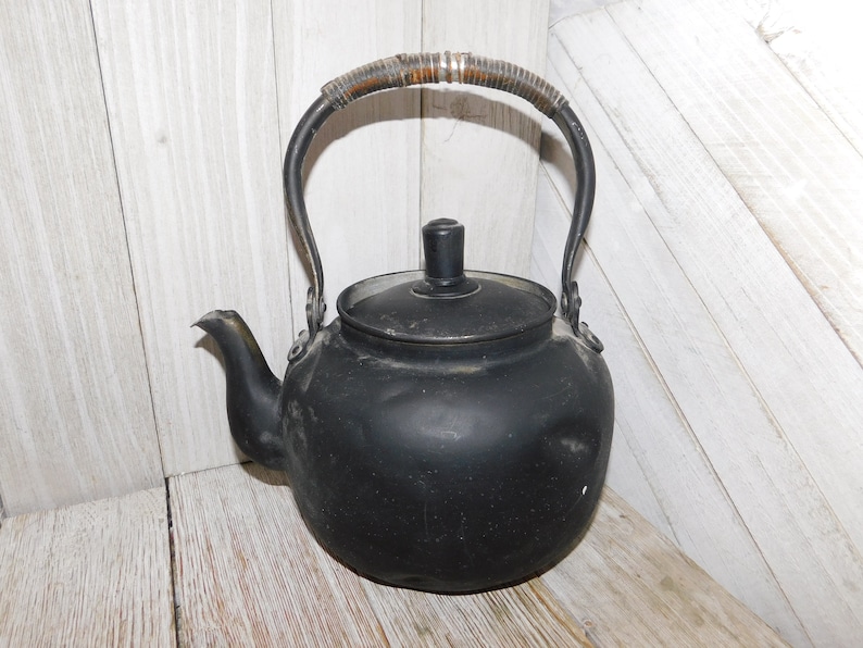 May include: A black metal teapot with a wooden handle. The teapot has a rounded body and a spout on the side. The teapot is sitting on a white wooden surface.