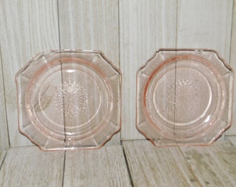 May include: Two pink glass plates with a scalloped edge and a central floral design. The plates have a square shape with rounded corners. The plates are displayed on a light wood surface.