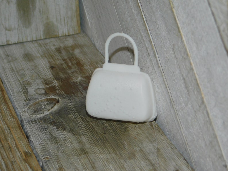 Op de afbeelding: Een klein, wit object in de vorm van een handtas met een handvat. De tas heeft een afgeronde rechthoekige vorm met een textuuroppervlak en een handvat. De tas ligt op een houten oppervlak.