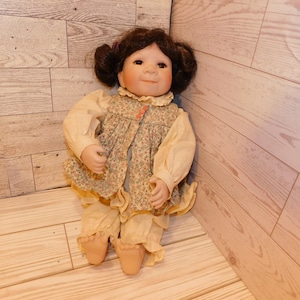 May include: A vintage porcelain doll with brown hair styled in two buns. The doll wears a floral print vest over a cream-colored blouse and matching ruffled pants. The doll is posed sitting on a wooden surface.