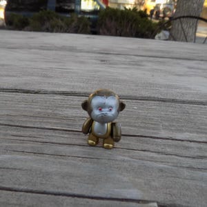 May include: A small, silver and gold monkey figurine with red eyes. The monkey is standing on a wooden surface.