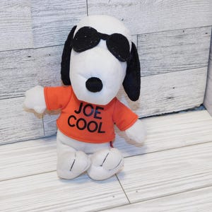 May include: A Snoopy plush toy wearing black sunglasses and an orange shirt with the text "JOE COOL". The plush has black ears, a black nose, and white body parts. The toy is sitting on a white wooden surface.