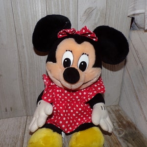 K&ouml;nnte beinhalten: Ein Pl&uuml;schtier von Minnie Mouse, die ein rotes Kleid mit wei&szlig;en Punkten und eine rote Schleife tr&auml;gt. Sie hat schwarze Ohren und eine schwarze Nase.