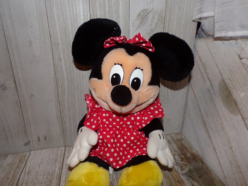 K&ouml;nnte beinhalten: Ein Pl&uuml;schtier von Minnie Mouse, die ein rotes Kleid mit wei&szlig;en Punkten und eine rote Schleife tr&auml;gt. Sie hat schwarze Ohren und eine schwarze Nase.