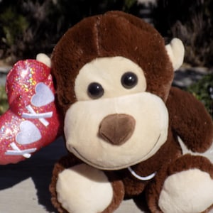 May include: A brown plush monkey toy holding a red heart-shaped balloon with white glitter and hearts. The monkey has a brown nose and brown eyes.
