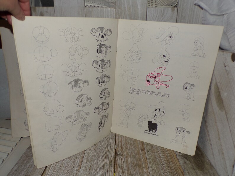 May include: Open book with pencil sketches of cartoon character faces. The sketches show the development of the faces, with step-by-step instructions. The text on the page reads, "Study the development of these expressions and work out some of your own."