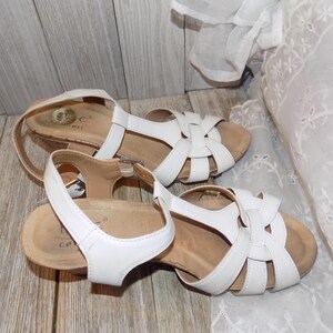 K&ouml;nnte beinhalten: Ein Paar wei&szlig;e Ledersandalen mit Korkkeil und einem Kreuzriemen-Design.