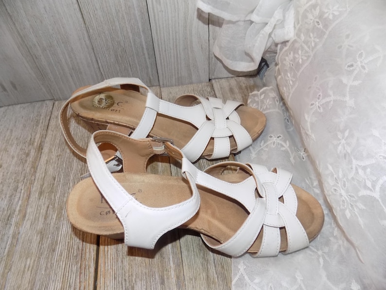K&ouml;nnte beinhalten: Ein Paar wei&szlig;e Ledersandalen mit Korkkeil und einem Kreuzriemen-Design.