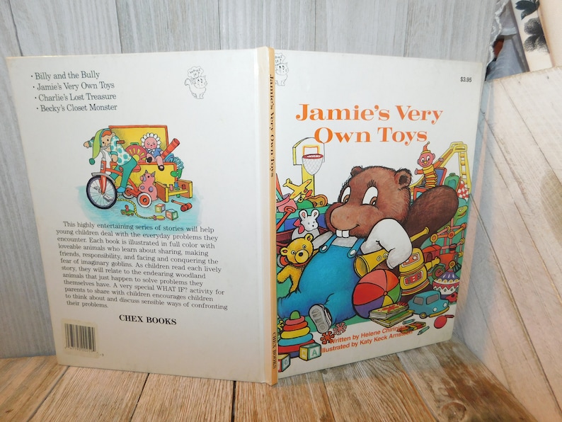 May include: A vintage children's book titled "Jamie's Very Own Toys" features a cartoon illustration of a beaver with a toothy grin, wearing overalls and surrounded by toys. The book cover is yellow and orange with a price tag of $2.95.