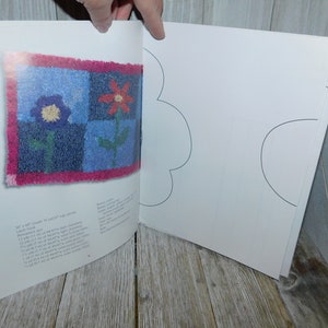 May include: A book titled "Bloom'n Happy" with a colorful rug design on the cover. The rug is made of squares with a red flower, a blue flower, and green leaves. The book is open to a page with a blank template for creating a rug.