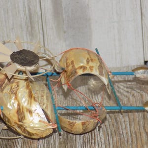 May include: A whimsical, handmade art piece featuring repurposed seashells and other natural materials. The sculpture incorporates a fan-like element, shell components, and blue and orange accents, creating a unique, textured design. The piece is set against a wooden background.
