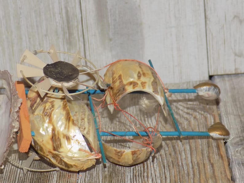 May include: A whimsical, handmade art piece featuring repurposed seashells and other natural materials. The sculpture incorporates a fan-like element, shell components, and blue and orange accents, creating a unique, textured design. The piece is set against a wooden background.
