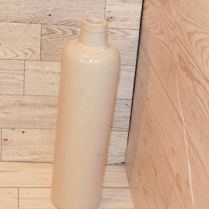 May include: A tall, cylindrical, off-white ceramic bottle with a narrow neck. The bottle has a smooth surface and a slightly rounded base. It is leaning against a wooden wall with a light brown color.