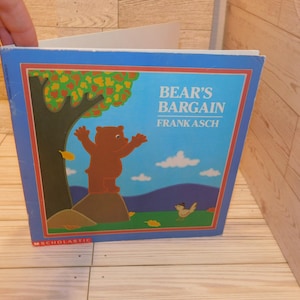 May include: A children's book titled "Bear's Bargain" by Frank Asch. The cover features a brown bear with outstretched arms on a rock, a tree with green and yellow leaves, and a small bird. The background is blue.
