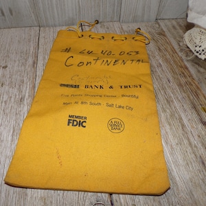 May include: Mustard-yellow canvas bag with a drawstring closure. The bag has handwritten text and printed text, including "Continental" and "Bank & Trust." The bag also has the text "Member FDIC" and "A Full Service Bank."