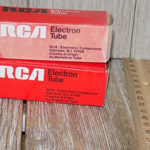 May include: Two red RCA Electron Tube boxes stacked on a wooden surface. The boxes have white text that reads "RCA Electron Tube" and include the address of the manufacturer. A wooden ruler is next to the boxes.