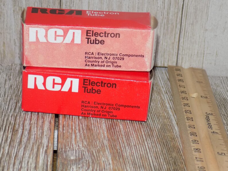 May include: Two red RCA Electron Tube boxes stacked on a wooden surface. The boxes have white text that reads "RCA Electron Tube" and include the address of the manufacturer. A wooden ruler is next to the boxes.