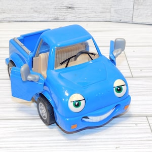 May include: A blue toy pickup truck with a smiling face, large eyes, and open doors. The truck has a tan interior, black steering wheel, and side mirrors. The tires are gray.