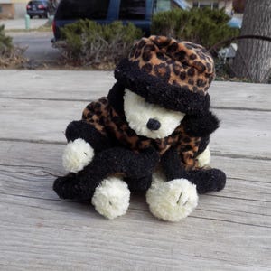 May include: A plush toy animal with a leopard print hat and coat, black trim, and white paws. The toy is sitting on a weathered wooden surface, with a neutral background of a street and trees.