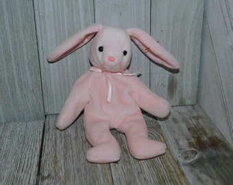 May include: A light pink plush rabbit toy with long ears, black plastic eyes, and a small pink nose. The stuffed animal features a thin pink ribbon tied in a bow around its neck and has a small red tag attached to the base of its left ear.