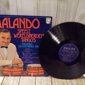 May include: Vintage vinyl record album titled "Malando Speelt Wereldbroem Tango's" featuring a portrait of a man in a suit. The album cover is blue with white and red text. A black vinyl record is next to the album.