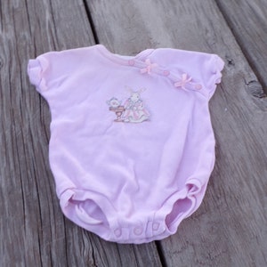 May include: A pink baby onesie with a white bunny and teacup illustration. The onesie has pink bows and buttons on the shoulder.