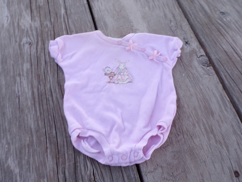 May include: A pink baby onesie with a white bunny and teacup illustration. The onesie has pink bows and buttons on the shoulder.