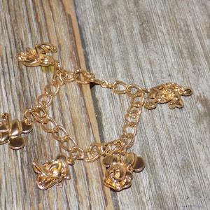 May include: Gold-coloured charm bracelet with Mickey Mouse, Donald Duck, and other character charms. The bracelet has a chain link design and is displayed on a wooden surface. The charms are detailed, adding a playful touch to the jewellery.