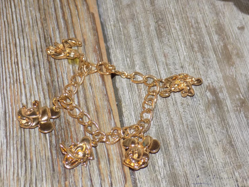 May include: Gold-coloured charm bracelet with Mickey Mouse, Donald Duck, and other character charms. The bracelet has a chain link design and is displayed on a wooden surface. The charms are detailed, adding a playful touch to the jewellery.