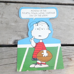 Peut inclure: Une illustration de bande dessin&eacute;e de Charlie Brown tenant un ballon de football sur un terrain vert. Le texte au-dessus de lui dit "MONDAY IS THE FOURTH-DOWN-LONG-YARDAGE DAY OF THE WEEK!"