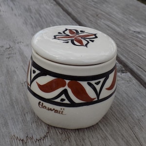 May include: A small, off-white ceramic jar with a lid. The jar is decorated with a black and brown geometric pattern and the word "Hawaii" is written on the front. The lid features a floral design in black and brown.