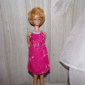 May include: A vintage Barbie doll with blonde hair wearing a pink dress with a blue trim and a floral pattern.