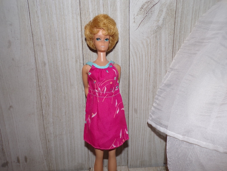 May include: A vintage Barbie doll with blonde hair wearing a pink dress with a blue trim and a floral pattern.