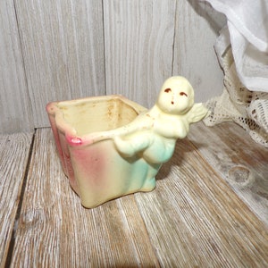 May include: A vintage ceramic planter shaped like a square with a cherub figure holding the planter. The planter is painted in shades of pink, yellow, and blue.
