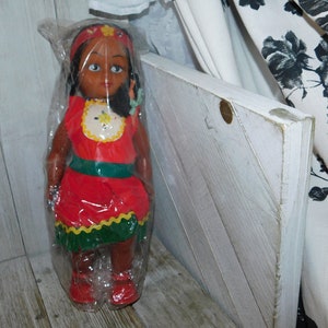 May include: A plastic doll with dark brown skin, black hair, and brown eyes. She is wearing a red dress with green and yellow trim. The doll is wrapped in plastic and is sitting on a wooden surface.