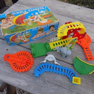 May include: A vintage toy train set called "Jungle Express" with a battery-operated engine, a green track, a yellow and red bridge, a gray elephant, and a white rhino. The set comes in a colorful box with illustrations of the train and animals.