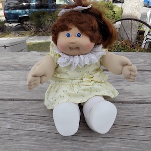 May include: A vintage doll with brown hair styled in a ponytail, blue eyes, and a soft, fabric body. The doll wears a yellow dress with a floral pattern and a white lace collar. White socks and shoes complete the outfit. The doll is sitting on a wooden surface.