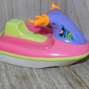 May include: A pink, green, and purple plastic toy jet ski with an orange handle. The jet ski has a sticker with a fish and the text "CPK".