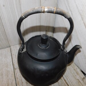 May include: Black cast iron kettle with a woven handle. The kettle has a rounded body and a lid with a knob.