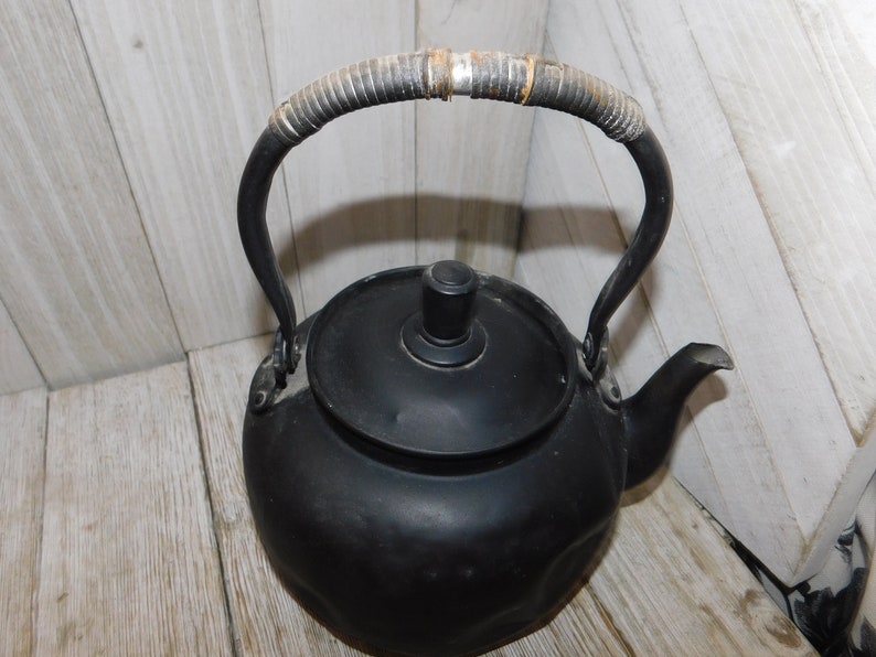 May include: Black cast iron kettle with a woven handle. The kettle has a rounded body and a lid with a knob.