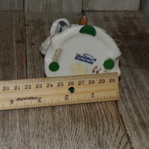 Pu&ograve; includere: Figurina in porcellana bianca di una donna con un vestito blu e bianco e un cappello verde. La statuetta &egrave; seduta su una base verde con il testo "Vonna-House" e "Blue Creek" sul fondo.