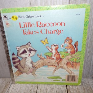 May include: A Little Golden Book titled "Little Raccoon Takes Charge". The cover features a raccoon sitting on a branch over a stream, with two chipmunks and a butterfly nearby. The book is blue with a green border.