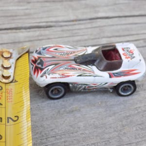 Puede incluir: Un coche de carreras de juguete blanco con gr&aacute;ficos tribales rojos, negros y plateados, una cabina abierta de color marr&oacute;n oscuro y ruedas negras. Una cinta m&eacute;trica amarilla est&aacute; junto al coche, mostrando una longitud de unos 7 cm.