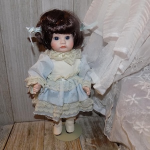 May include: A porcelain doll with brown hair styled in pigtails, decorated with blue ribbons. The doll is dressed in a light blue dress with white lace trim and white shoes. The doll stands on a small, round, beige stand.