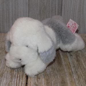 May include: A white and gray plush toy dog with a floppy ear. The dog is lying on its side with its head resting on its paw. The toy has a pink tag attached to it with the text "CE".