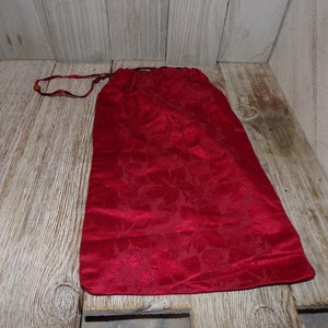 May include: A deep red drawstring bag made of a velvety fabric with a floral pattern. The bag is closed with a matching red drawstring. The bag is resting on a wooden surface.